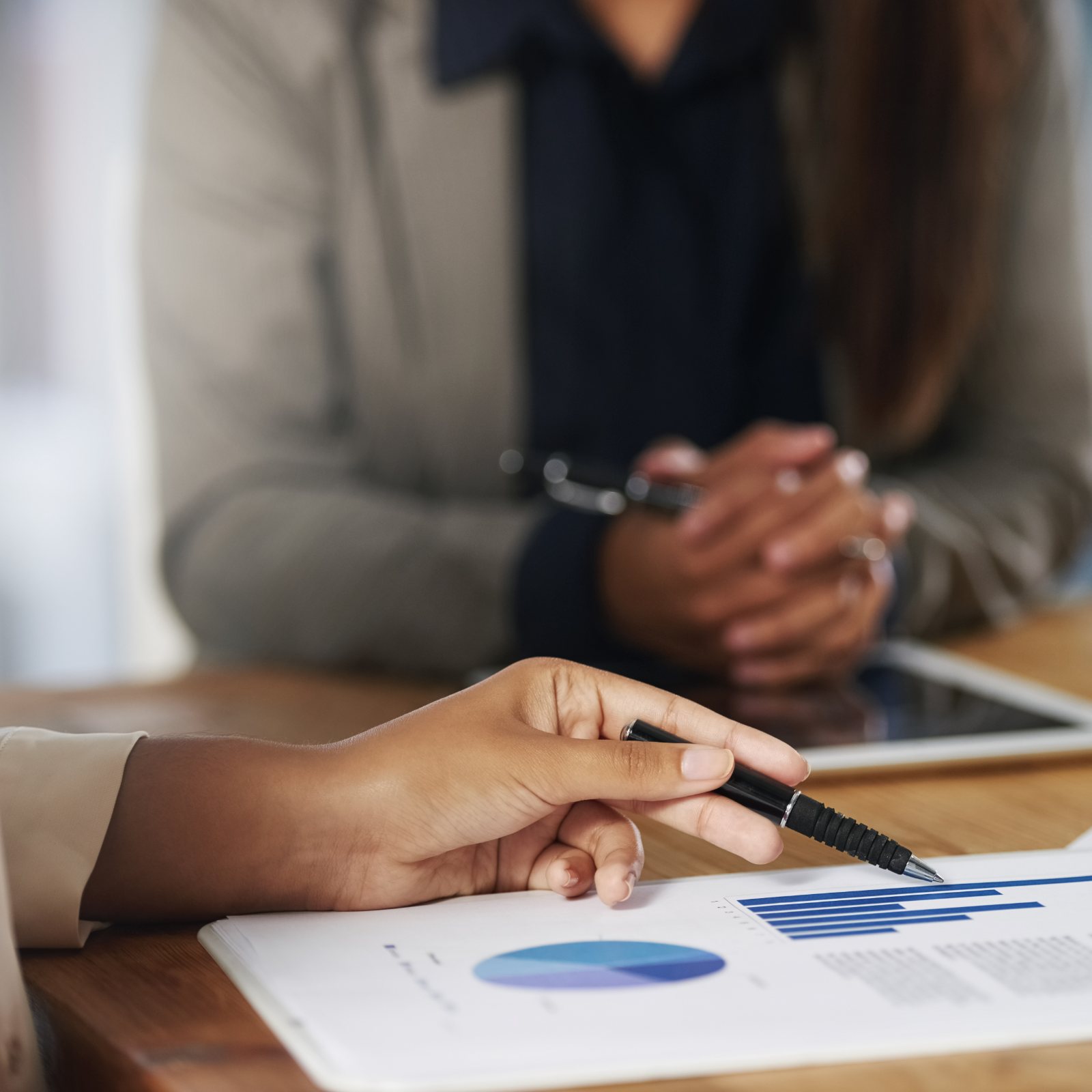 Two people at a desk, a hand with a pen pointing at a printed bar chart and pie chart